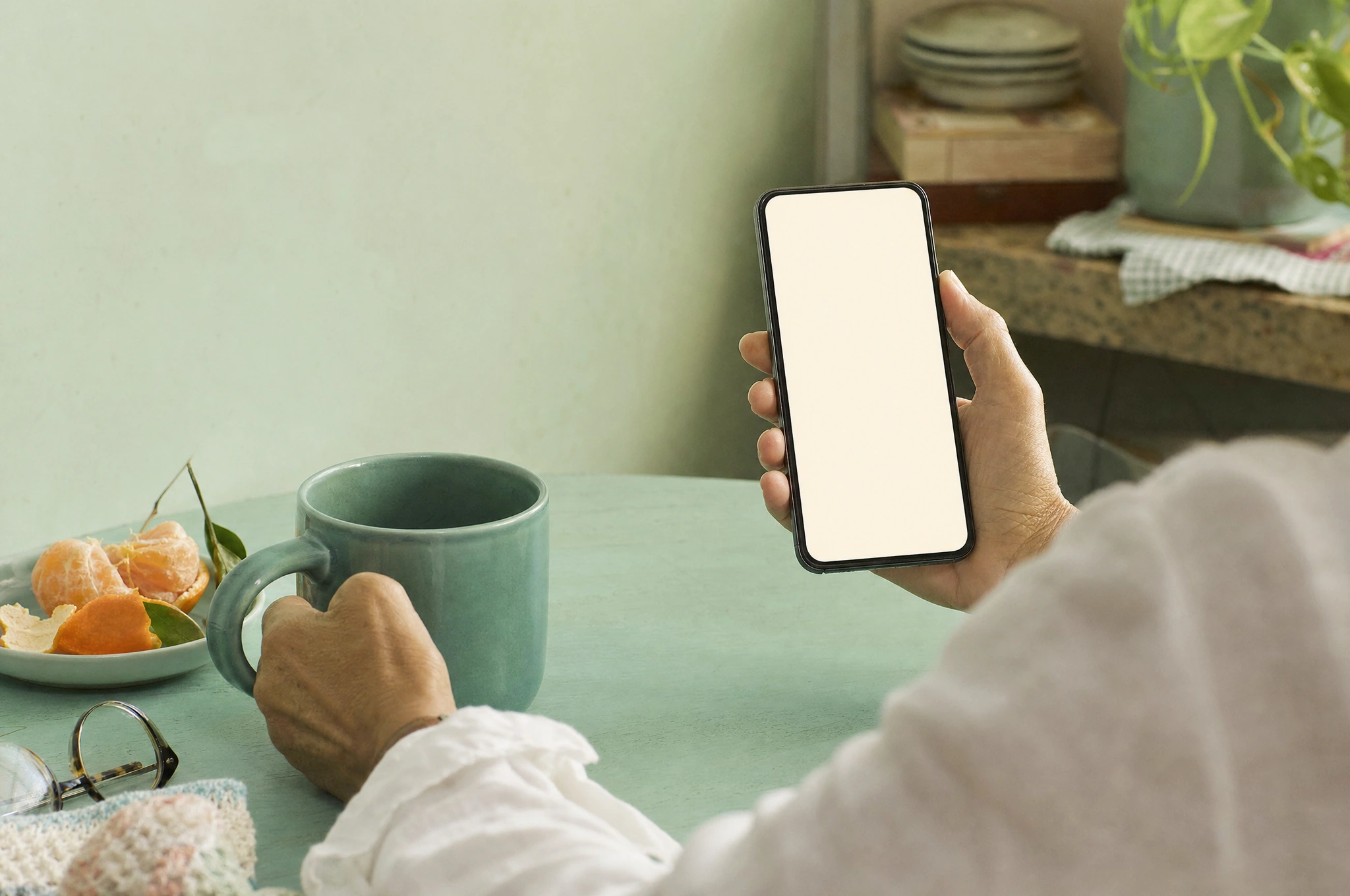 Hands with a phone and teal mug at a mint kitchen table in warm morning light